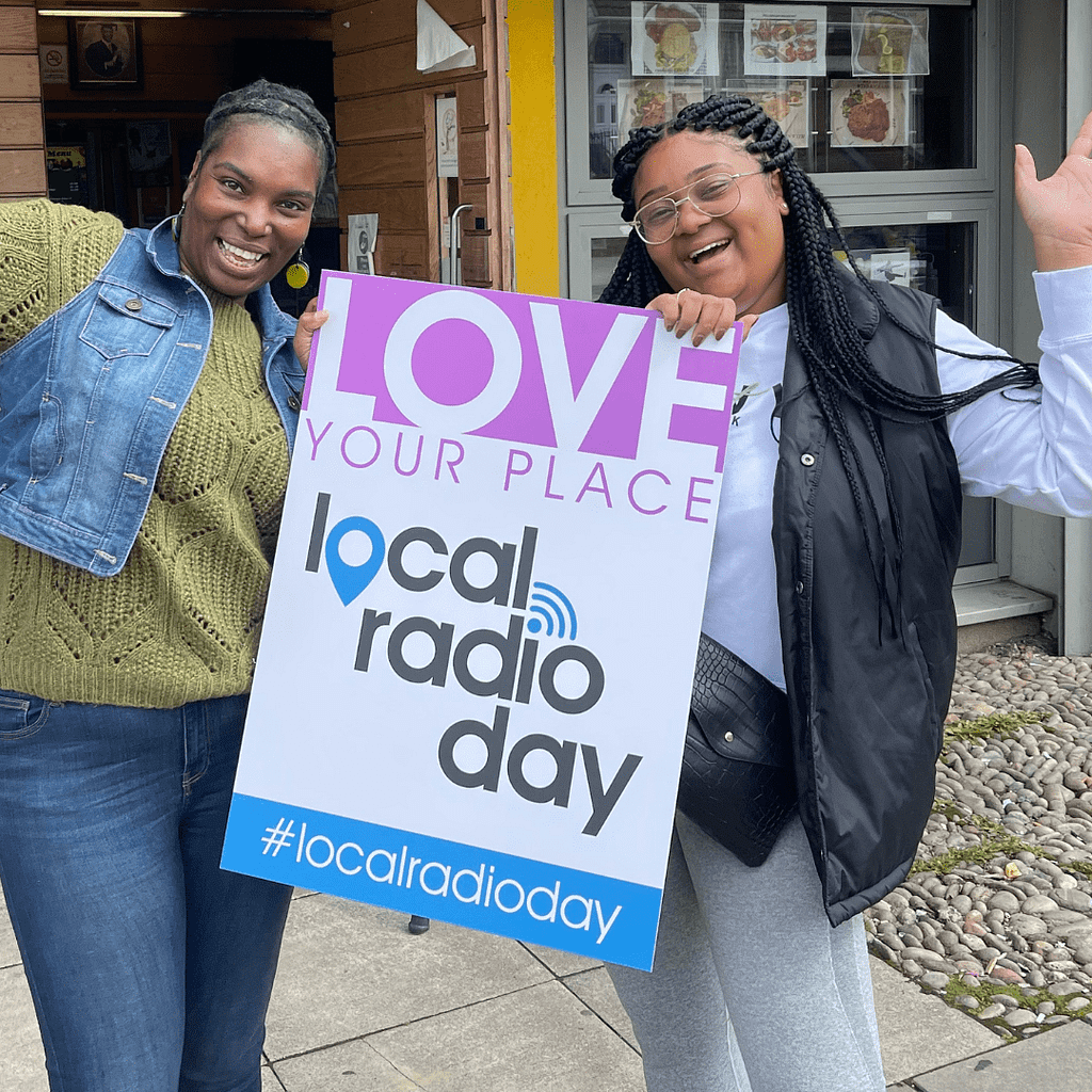 Image of two female radio station presenters celeberating Local Radio in 2021. They are holding a sign which shares the theme 'Love your place'.